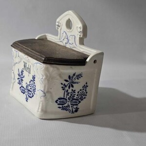 May include: White ceramic wall-mounted container with a dark wooden lid. The container is decorated with blue floral patterns and the word "Mehl". It has a small handle with a hole for hanging.