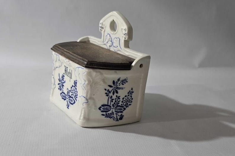 May include: White ceramic wall-mounted container with a dark wooden lid. The container is decorated with blue floral patterns and the word "Mehl". It has a small handle with a hole for hanging.