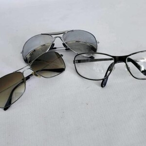 Puede incluir: Tres pares de gafas se muestran sobre una superficie blanca. Dos pares de gafas de sol tienen monturas met&aacute;licas y lentes tintadas, una con acabado de espejo. El tercer par son gafas con montura negra y lentes transparentes.