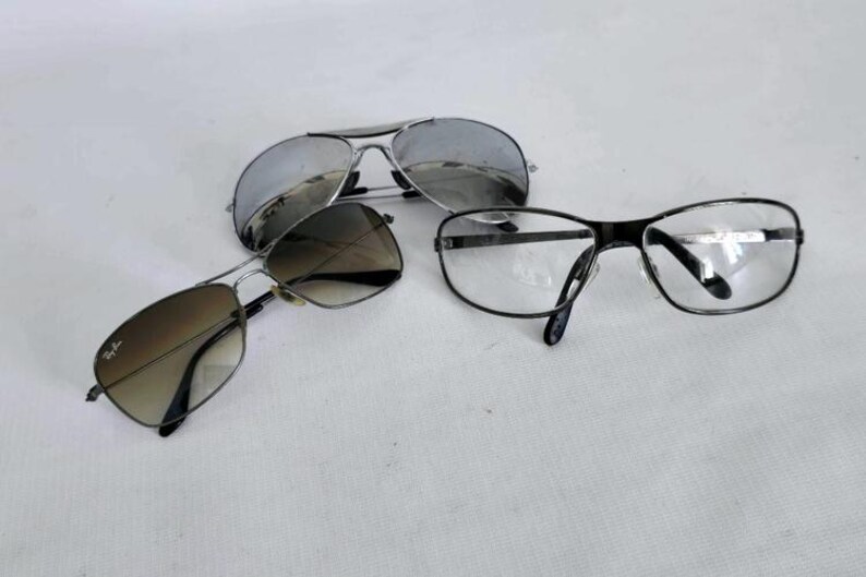 Puede incluir: Tres pares de gafas se muestran sobre una superficie blanca. Dos pares de gafas de sol tienen monturas met&aacute;licas y lentes tintadas, una con acabado de espejo. El tercer par son gafas con montura negra y lentes transparentes.