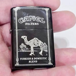 May include: Black lighter with white text and a camel graphic. The text on the lighter reads "CAMEL FILTERS" and "TURKISH & DOMESTIC BLEND". The lighter has a rectangular shape and a hinged lid.