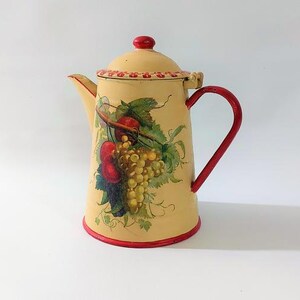 May include: Cream-coloured vintage enamelware coffee pot with a red handle and trim. The pot features a painted design of grapes and apples. The lid and handle are red, and the body is a light cream colour.