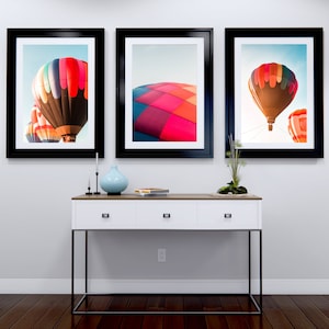 Può includere: Tre immagini incorniciate di mongolfiere su uno sfondo di cielo blu. Le immagini sono appese a una parete bianca sopra un tavolo consolle bianco con gambe in metallo nero.