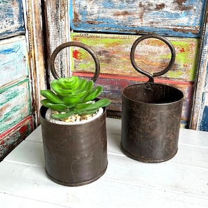 May include: Two cylindrical dark brown metal containers with circular top handles. One container holds a small green artificial succulent plant nestled in light-coloured pebbles. The metal surfaces feature a weathered, rustic texture throughout.