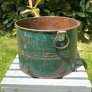 May include: A cylindrical green metal bucket featuring a weathered finish with visible rust patches, a prominent metal ring handle attached to the side, and the white painted text AN-437 on the exterior surface of the container.