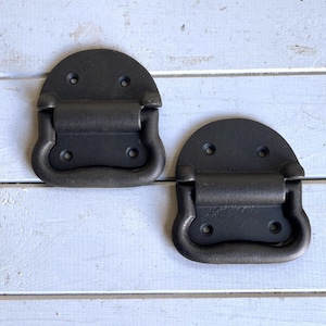 May include: Two dark grey metal chest handles with a semi-circular mounting plate and a hinged drop-down pull. Each handle features four pre-drilled screw holes for attachment, showcasing a matte finish and a sturdy, functional design.