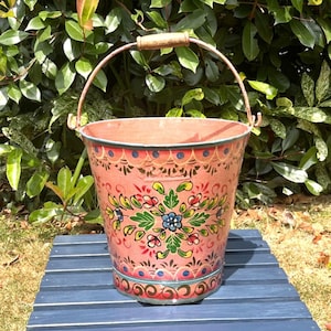 May include: A tapered metal bucket with a pink base colour, featuring a hand-painted floral pattern in green, blue, red, and yellow. The bucket has a metal handle with a wooden grip at the top and a decorative scalloped border design.