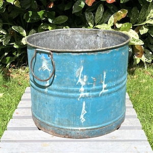 May include: A cylindrical metal bucket featuring a distressed bright blue painted exterior with visible white paint streaks and signs of wear. The container has a wide open top, a metallic rim, and a single curved metal handle attached to the side.