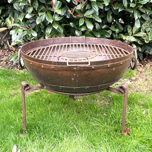 May include: A round, rusted metal fire pit bowl featuring a riveted rim, two side handles, and a removable circular grill grate insert. The bowl is supported by three thin, curved metal legs that extend outward from the base of the structure.