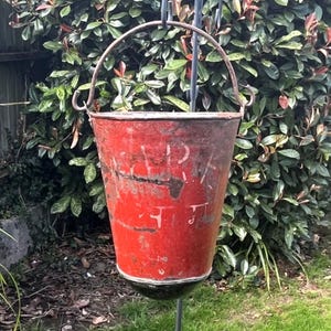 May include: A vintage red metal fire bucket with a weathered finish and a curved handle. The exterior surface shows signs of wear, including rust and chipped paint, with faint, partially obscured white lettering visible on the front side.