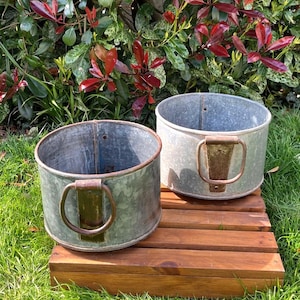 May include: Two round galvanised metal planters with weathered surfaces and prominent, rusted iron ring handles attached to the sides. These cylindrical containers feature a metallic grey finish with visible signs of oxidation and wear on the hardware.