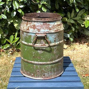 May include: A cylindrical metal milk churn featuring a weathered finish with patches of green and light blue paint. The container displays visible rust, a wide open top, and a sturdy metal handle attached to the side for lifting and transport.