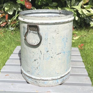 May include: A cylindrical metal bucket with a distressed light blue and white painted finish. The bucket features a dark, rusted metal drop handle attached to the side near the top rim. The interior of the bucket shows a raw, unpainted galvanised metal surface.