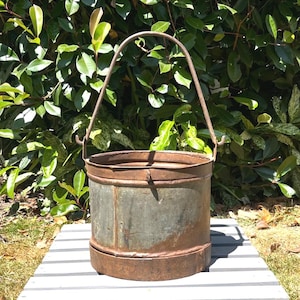 May include: A cylindrical metal bucket featuring a weathered, oxidised finish with visible rust patches along the base and rim. The container includes a tall, arched metal handle attached to two side hooks, providing a sturdy grip for transport.