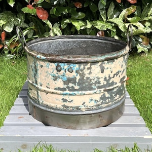 May include: A round metal basin featuring a distressed finish with layers of chipped cream and blue paint over a dark grey base. The vessel includes two curved metal handles on opposite sides and a smooth, unpainted metal band around the base.