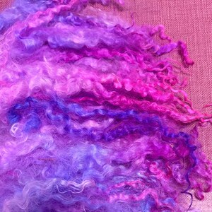 May include: Close-up of colorful fiber art material in shades of purple, pink, and blue. The fiber has a curly texture and is arranged in a layered pattern. The background is a textured pink surface.