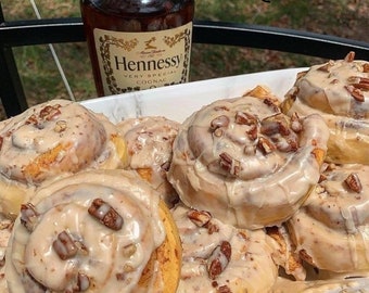 May include: A plate of cinnamon rolls topped with a white icing and pecan pieces. A bottle of Hennessy Very Special Cognac is in the background.