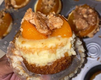 May include: A close-up of a peach cobbler cheesecake cupcake with a crumb topping. The cupcake is in a paper liner and is being held by a hand.
