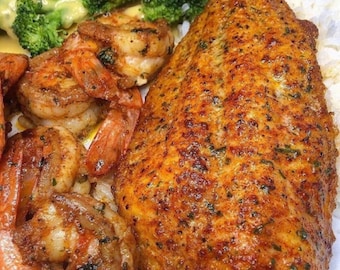 Puede incluir: Un plato de comida con un trozo de pescado frito, camarones, arroz y brócoli. El pescado está sazonado con hierbas y especias y tiene una corteza crujiente. Los camarones también están sazonados y cocinados. El arroz es blanco y esponjoso. El brócoli está al vapor y tiene un color verde brillante.