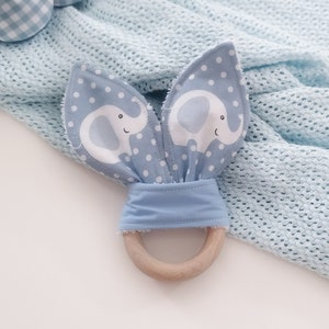 May include: A blue and white baby teether with elephant ears and a wooden ring. The teether is on a light blue blanket with a white background.