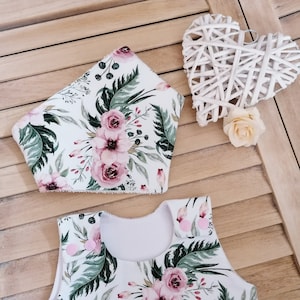 May include: A white sleeveless dress with a pink and green floral print. The dress has a ruffled hem and pink buttons on the straps. A matching pink and green floral print bib is included.
