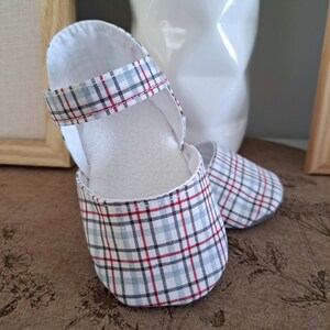 May include: A pair of baby shoes with a plaid pattern in red, blue, and white. The shoes have a Mary Jane style with a strap and a soft, white interior. The shoes are made of fabric and are designed for infants.
