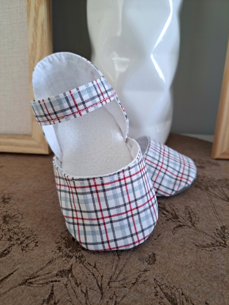 May include: A pair of baby shoes with a plaid pattern in red, blue, and white. The shoes have a Mary Jane style with a strap and a soft, white interior. The shoes are made of fabric and are designed for infants.