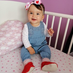 May include: A baby girl wearing a blue denim overall dress with white polka dots and a pink and red heart headband. She is sitting on a white crib mattress with pink stars. The baby is wearing red shoes with white soles.