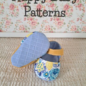 May include: Baby shoes with a floral pattern in blue and yellow, featuring a quilted light blue sole and a mustard yellow strap. The background includes the text "Happy Baby Patterns" over a floral design.