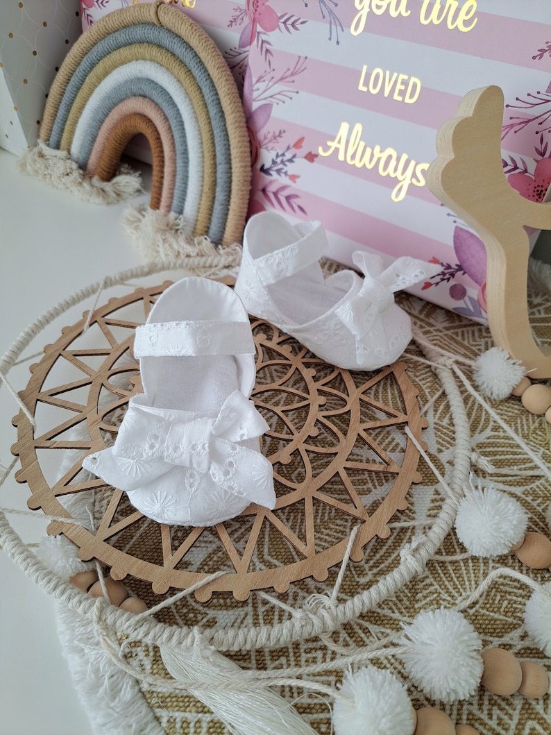 May include: White baby shoes with delicate floral embroidery and a bow, alongside a matching headband. The items are displayed on a wooden dreamcatcher with a macrame design. A decorative rainbow and a pillow with the words "you are LOVED Always" are in the background.