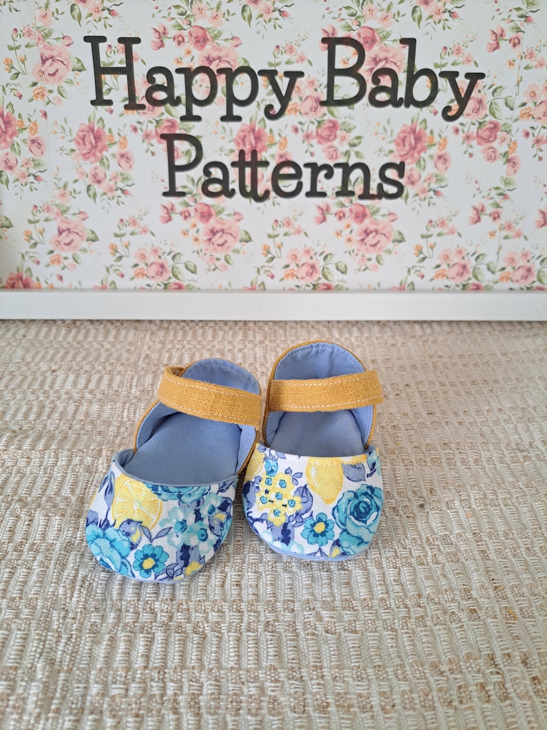 May include: A pair of baby shoes with a floral lemon pattern on the toe and a light blue interior. The shoes have a mustard yellow strap and are set against a floral background with the text "Happy Baby Patterns".