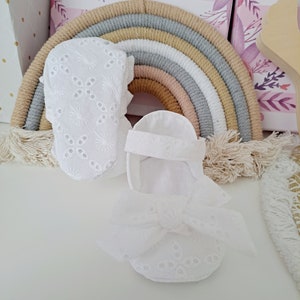 May include: White baby booties and sandals with eyelet embroidery and bow details. The shoes are set against a backdrop of a rainbow-shaped wall hanging and decorative boxes, suggesting a nursery or baby-themed setting.