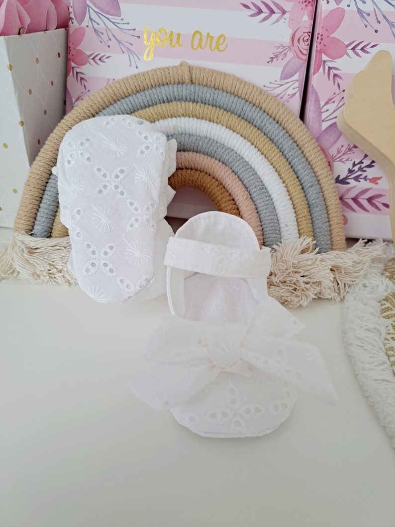May include: White baby booties and sandals with eyelet embroidery and bow details. The shoes are set against a backdrop of a rainbow-shaped wall hanging and decorative boxes, suggesting a nursery or baby-themed setting.
