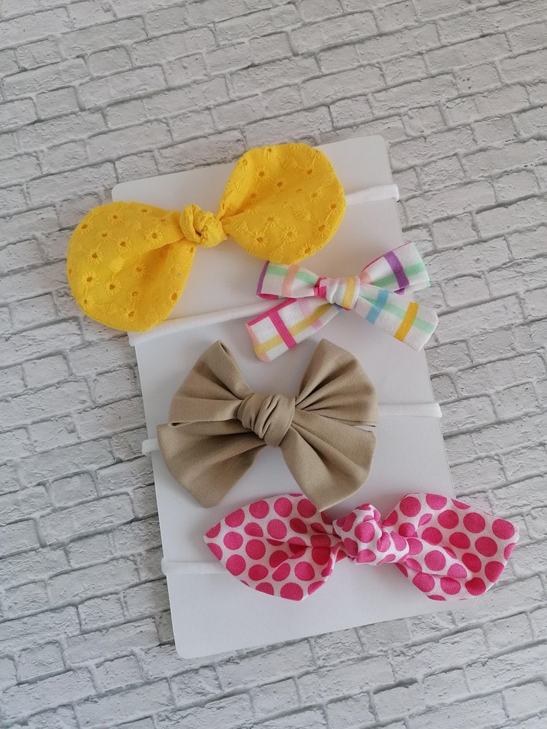 May include: Set of four hair bows on a white card. The bows are made of different fabrics, including yellow, pink, brown, and multicolored plaid. The bows are tied with a knot in the center.