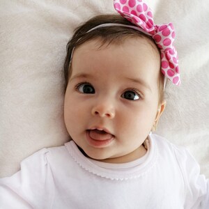 May include: A baby girl wearing a white long-sleeved shirt with a white lace trim around the neckline. She is wearing a pink headband with white polka dots and has a big smile on her face.