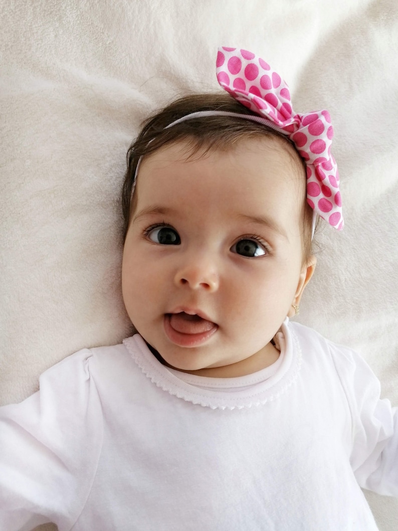 May include: A baby girl wearing a white long-sleeved shirt with a white lace trim around the neckline. She is wearing a pink headband with white polka dots and has a big smile on her face.
