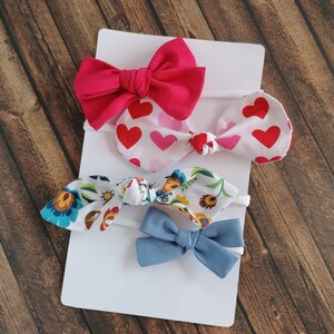May include: Set of four hair bows in various colors and patterns. The set includes a pink bow, a white bow with red hearts, a white bow with floral print, and a blue bow.