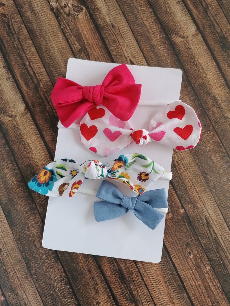 May include: Set of four hair bows in various colors and patterns. The set includes a pink bow, a white bow with red hearts, a white bow with floral print, and a blue bow.