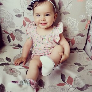May include: A baby girl wearing a pink floral dress with a white and blue striped headband and white floral booties sits on a floral patterned couch.