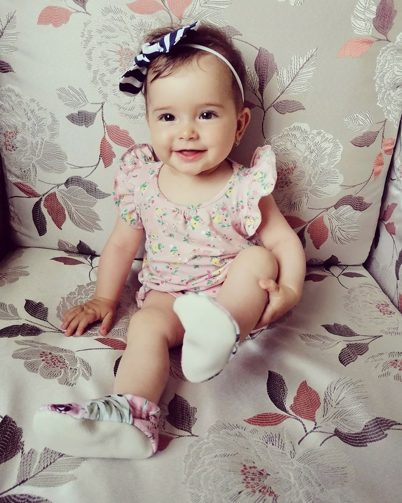 May include: A baby girl wearing a pink floral dress with a white and blue striped headband and white floral booties sits on a floral patterned couch.