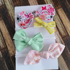 May include: Set of four hair bows with different patterns. The bows are made of fabric and are attached to elastic headbands. The patterns include floral, polka dot, and striped.