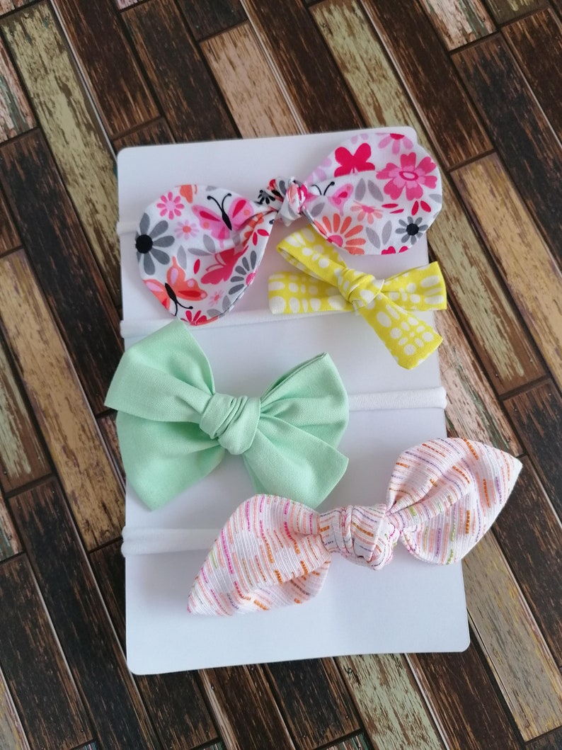 May include: Set of four hair bows with different patterns. The bows are made of fabric and are attached to elastic headbands. The patterns include floral, polka dot, and striped.
