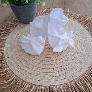 May include: A pair of white baby shoes with eyelet detailing and bow accents. The shoes are made of a soft fabric and are designed with a Mary Jane style. They are placed on a woven placemat with a fringed edge.