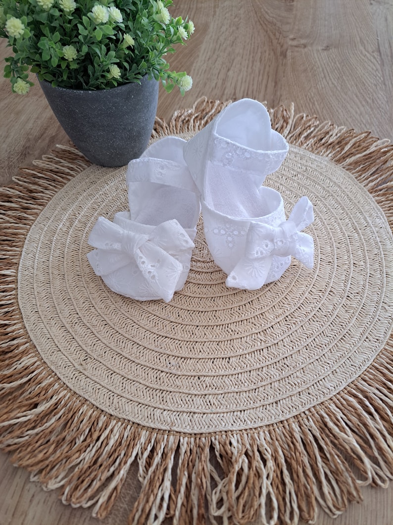 May include: A pair of white baby shoes with eyelet detailing and bow accents. The shoes are made of a soft fabric and are designed with a Mary Jane style. They are placed on a woven placemat with a fringed edge.