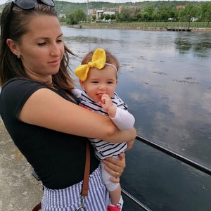May include: A woman in a black shirt and blue and white striped pants holds a baby wearing a striped shirt and a yellow bow. The baby is wearing watermelon-patterned shoes. The woman is holding a brown leather bag with a strap.