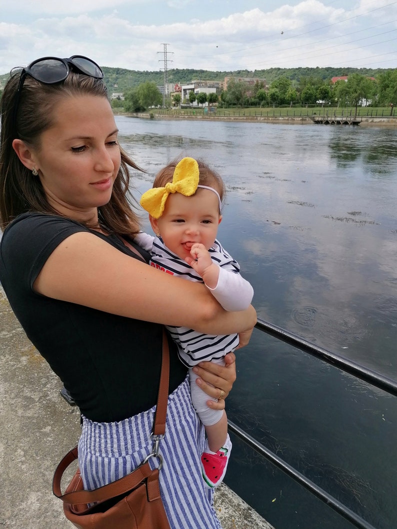 May include: A woman in a black shirt and blue and white striped pants holds a baby wearing a striped shirt and a yellow bow. The baby is wearing watermelon-patterned shoes. The woman is holding a brown leather bag with a strap.