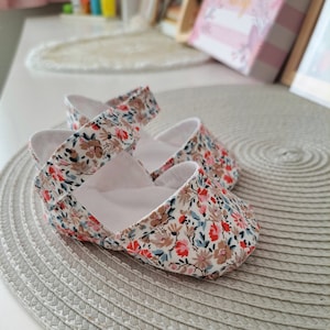 May include: A pair of baby shoes with a floral pattern in red, pink, brown, and blue on a white background. The shoes have a strap across the top and are made of soft fabric. The shoes are on a gray textured surface.