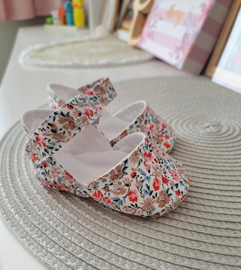 May include: A pair of baby shoes with a floral pattern in red, pink, brown, and blue on a white background. The shoes have a strap across the top and are made of soft fabric. The shoes are on a gray textured surface.
