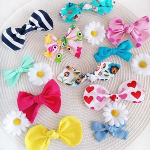 May include: A collection of colorful fabric hair bows with various patterns, including stripes, polka dots, hearts, and floral designs. The bows are arranged on a woven surface with white daisies.