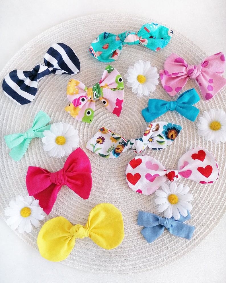 May include: A collection of colorful fabric hair bows with various patterns, including stripes, polka dots, hearts, and floral designs. The bows are arranged on a woven surface with white daisies.